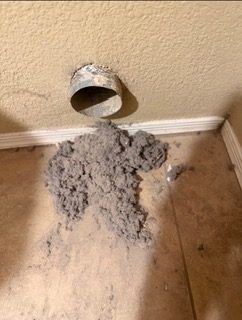 A pile of lint on the floor under a wall dryer vent, pulled out of one line
